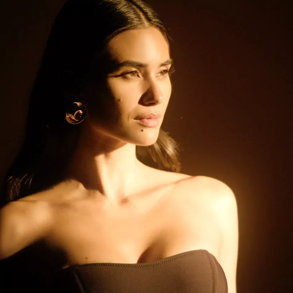 A person with long brunette hair poses in warm afternoon sunlight against a brown background, showing a confident expression.