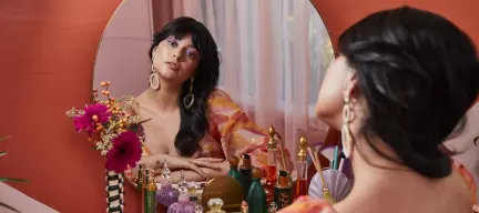 A person with long dark hair and colorful eye makeup sits in front of a vanity mirror adorned with various cosmetics and a bouquet of flowers.