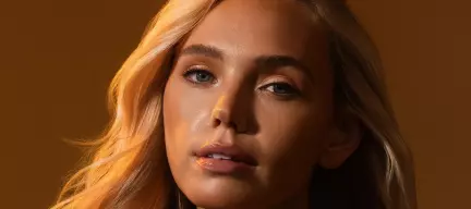 Close-up of a blonde woman with smooth, glowing skin and subtle makeup, lit to highlight her facial features against a neutral background.