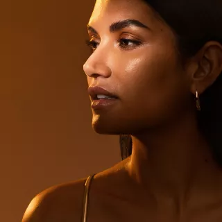 Close‑up profile of a woman with dark hair and glowing skin looking to the left against a warm brown background.