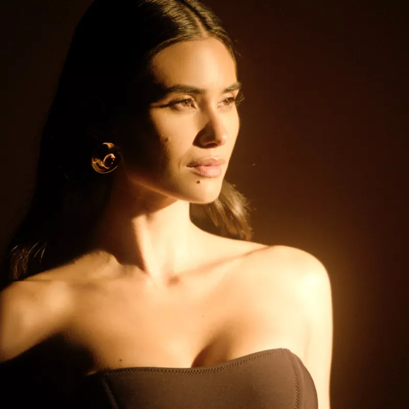 A person with long brunette hair poses in warm afternoon sunlight against a brown background, showing a confident expression.