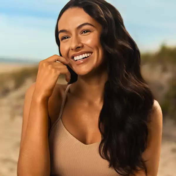 Woman with long dark hair wearing a white top, smiling and looking slightly to the side against a neutral background.