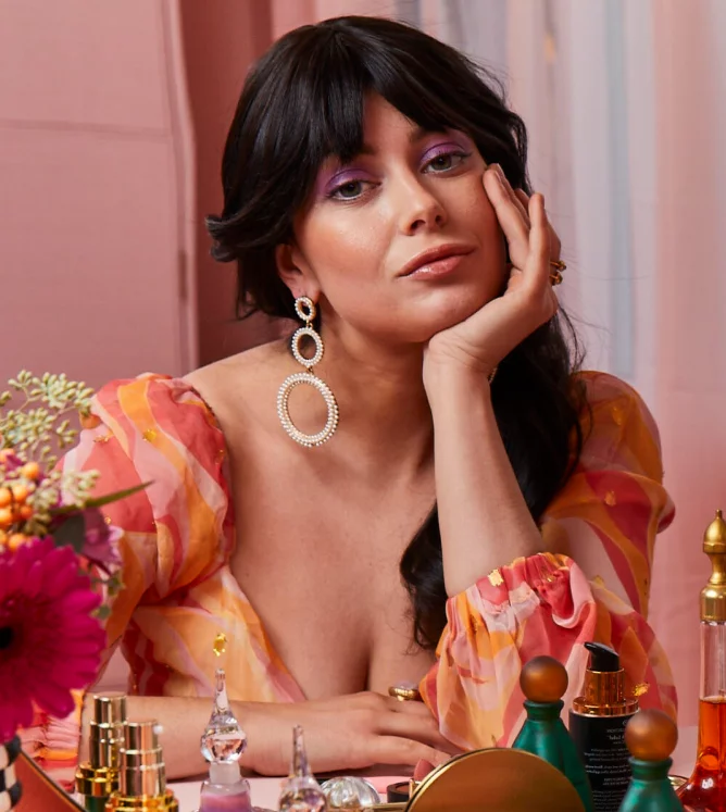 A female presenting person with dark hair and bangs wears a floral top and sits at a vanity