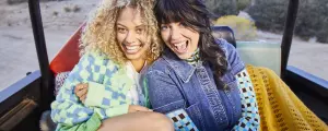 Two women sit in the open back of a Jeep, parked on a beach, laughing