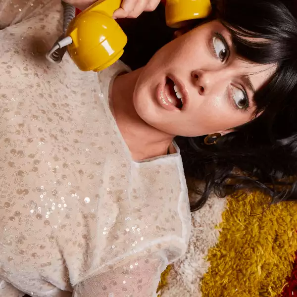 Woman with dark hair and bangs wearing a sparkly top holds a yellow retro-style telephone receiver near her face