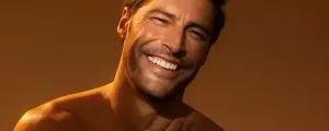 Close‑up of a shirtless man smiling and touching his shoulder with warm lighting.