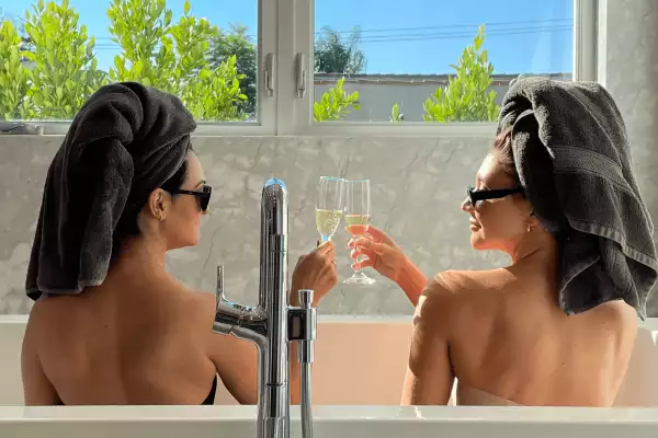 Two women with towels wrapping their hair and sunglasses clink classes while sitting in a large bathtub.