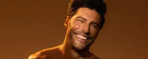 Smiling shirtless man with dark hair against a warm brown background.