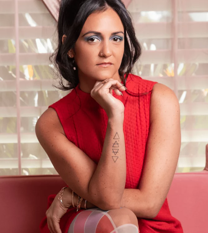 A female presenting person wearing a red sleeveless top rests her chin on her hand and stares directly into the camera
