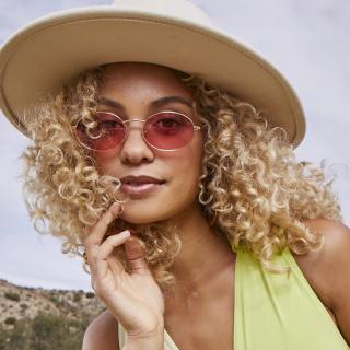 A woman with curly blonde hair stands in a desert landscape. She is wearing sunglasses, a large hat and a lime-green jumpsuit.