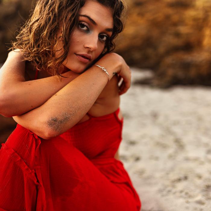 A woman in a red dress sits on a beach and stares into the camera.